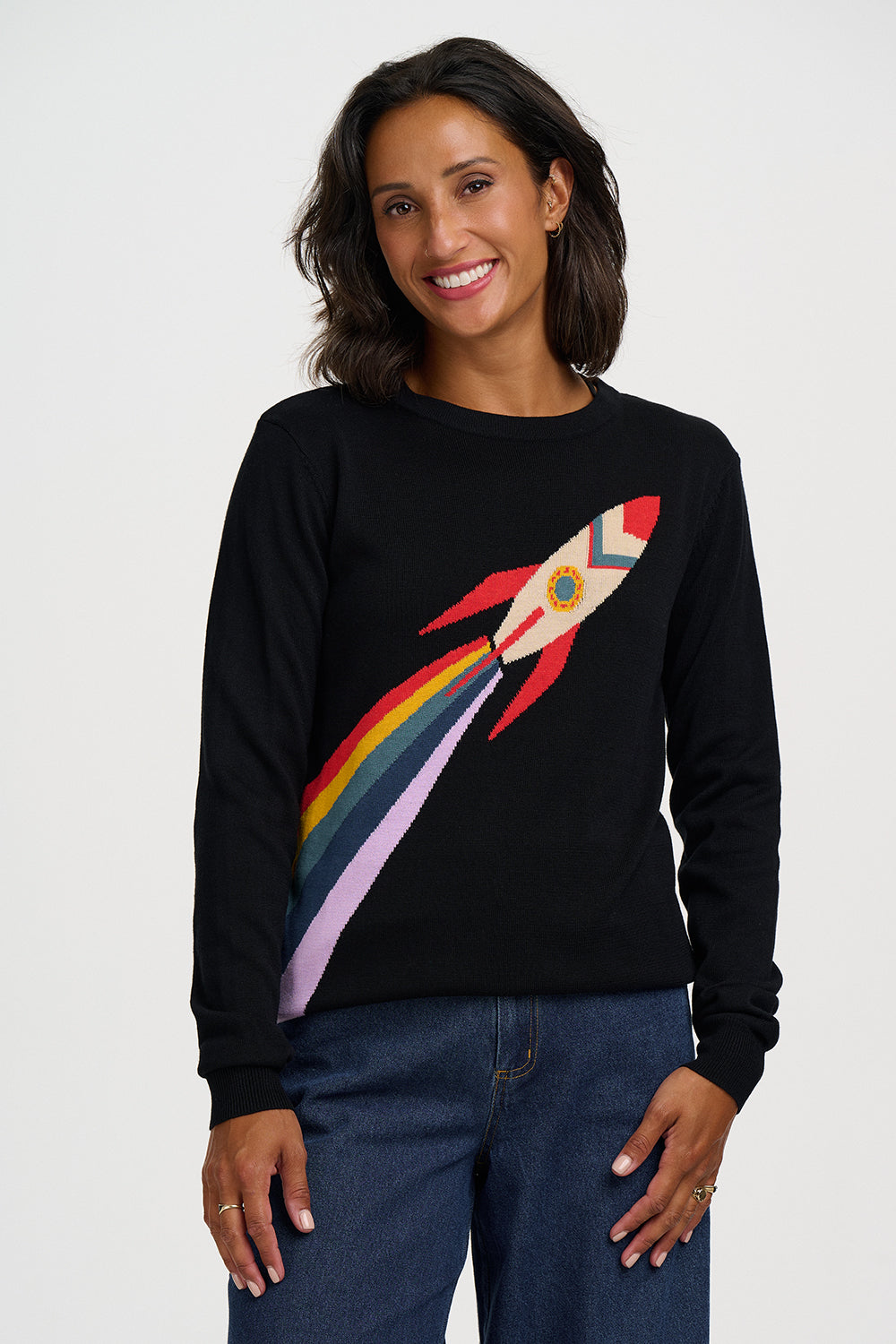 A woman wearing a black crewneck sweatshirt with a colorful rocket ship graphic on the front, paired with blue jeans, against a plain light gray background.