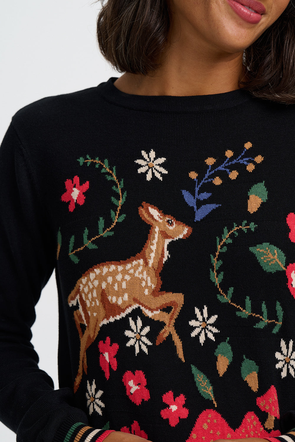 A close-up of a woman wearing a black crewneck sweater with a colorful knit design featuring a brown deer, red flowers, green leaves, and white daisies.
