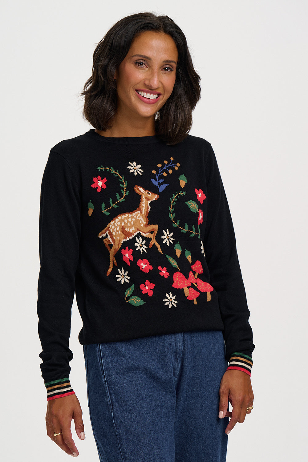 A woman wearing a black crewneck sweatshirt with a colorful embroidered deer and floral design, paired with blue jeans, against a light gray studio background.