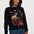 A woman wearing a black crewneck sweatshirt with a colorful embroidered deer and floral design, paired with blue jeans, against a light gray studio background.