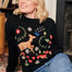 A woman sitting on a patterned floor wearing a black jumper with a colorful woodland animal and floral design, paired with blue jeans.