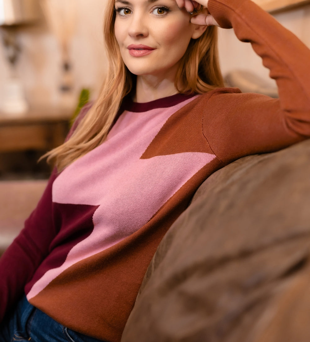 A woman with red hair wearing a color-blocked knit sweater with brown, dusty pink, and burgundy panels, sitting on a couch in a warm, softly lit living room.
