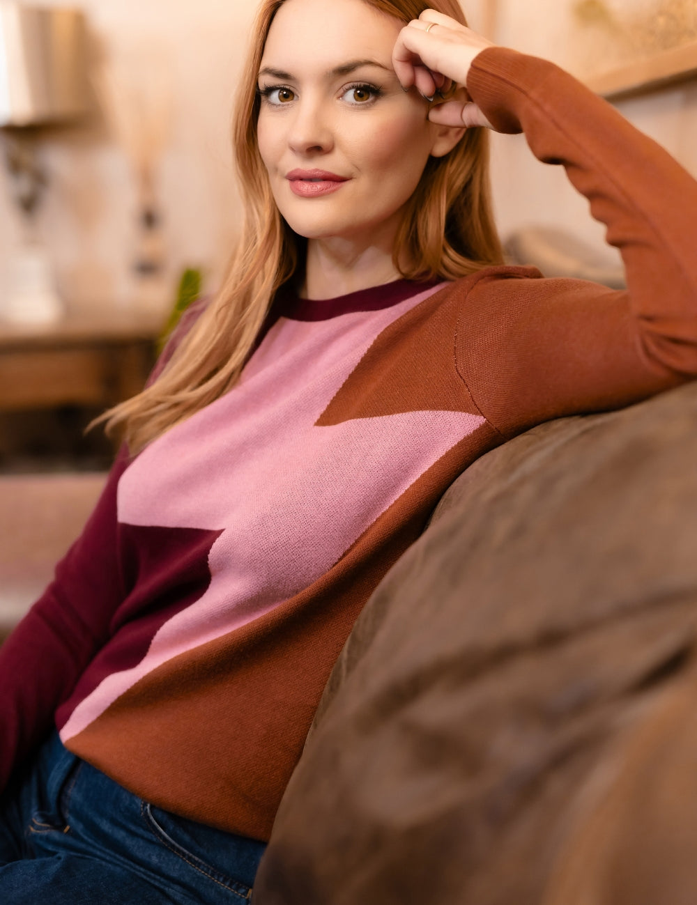 A woman with red hair wearing a color-blocked knit sweater with brown, dusty pink, and burgundy panels, sitting on a couch in a warm, softly lit living room.