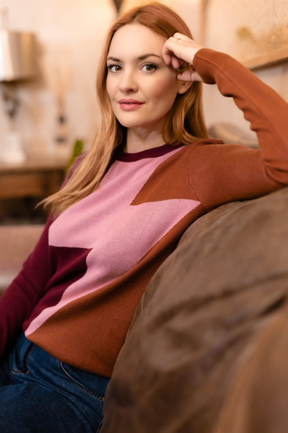 A woman with red hair wearing a color-blocked knit sweater with brown, dusty pink, and burgundy panels, sitting on a couch in a warm, softly lit living room.