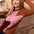 A woman with red hair wearing a color-blocked knit sweater with brown, dusty pink, and burgundy panels, sitting on a couch in a warm, softly lit living room.