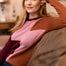 A woman with red hair wearing a color-blocked knit sweater with brown, dusty pink, and burgundy panels, sitting on a couch in a warm, softly lit living room.