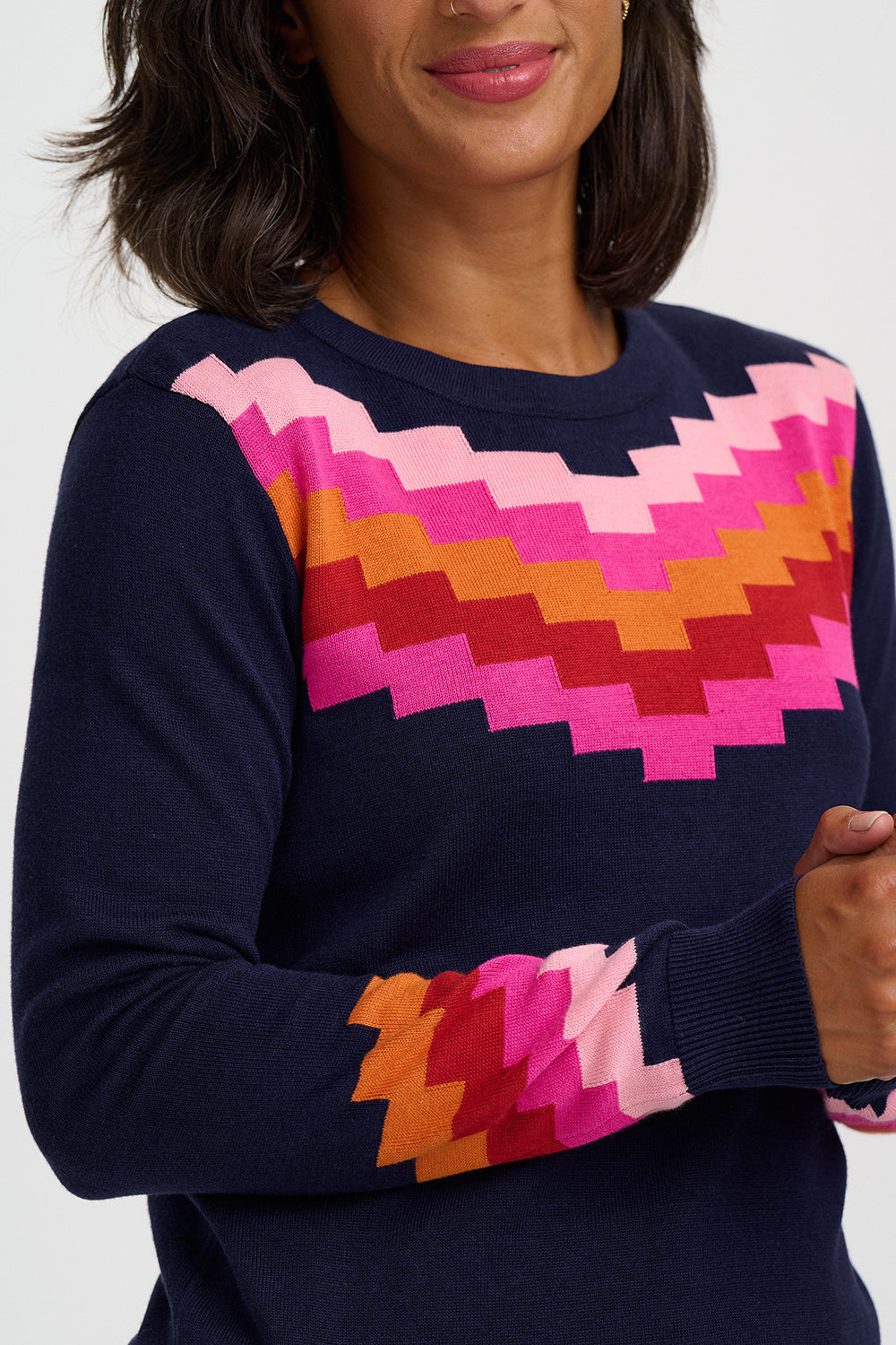 A close-up of a woman wearing a navy blue crewneck sweater with a colorful geometric chevron pattern in pink, orange, and red across the chest and sleeve.