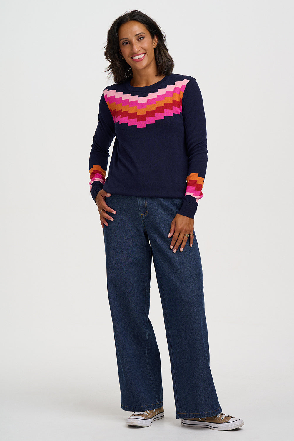 A woman modeling a navy blue knit sweater with a colorful geometric pattern across the chest and cuffs, paired with dark blue jeans and brown sneakers, against a light gray studio background.