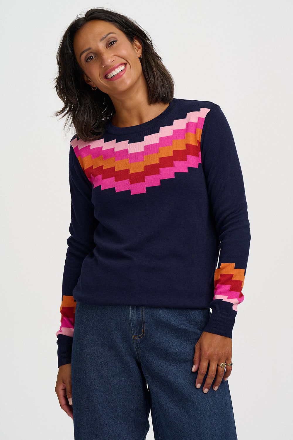 A woman wearing a navy blue crewneck sweater with a colorful geometric pixelated pattern across the chest and cuffs, paired with blue jeans, against a plain light gray background.