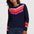 A woman wearing a navy blue crewneck sweater with a colorful geometric pixelated pattern across the chest and cuffs, paired with blue jeans, against a plain light gray background.