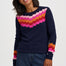 A woman wearing a navy blue crewneck sweater with a colorful geometric pixelated pattern across the chest and cuffs, paired with blue jeans, against a plain light gray background.