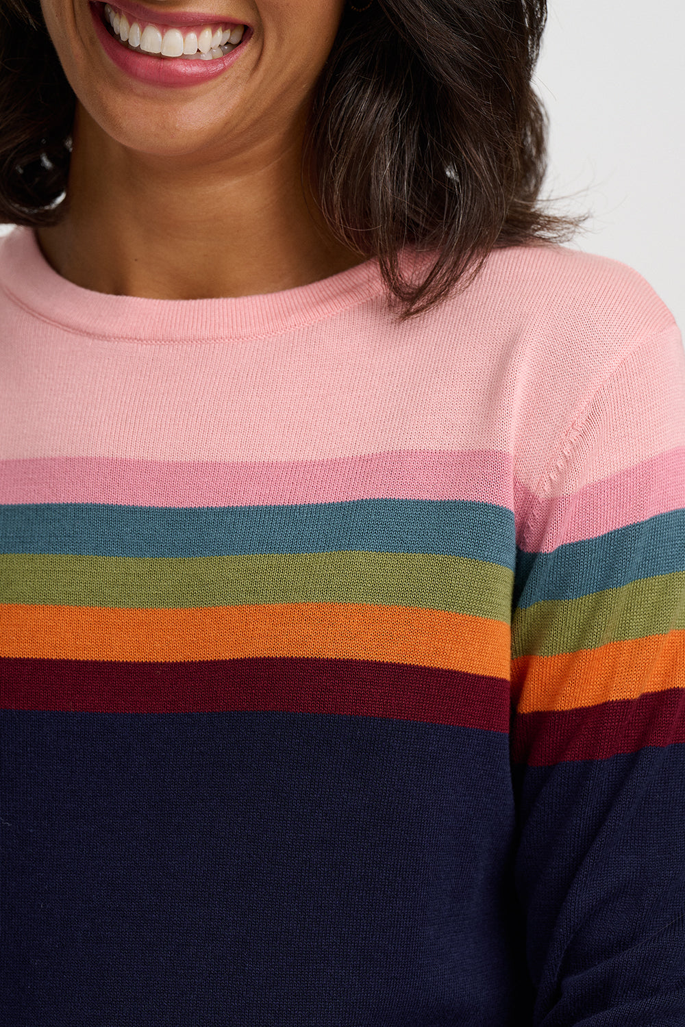A close-up of a woman wearing a crewneck sweater with horizontal stripes in pink, teal, olive green, orange, burgundy, and navy blue.