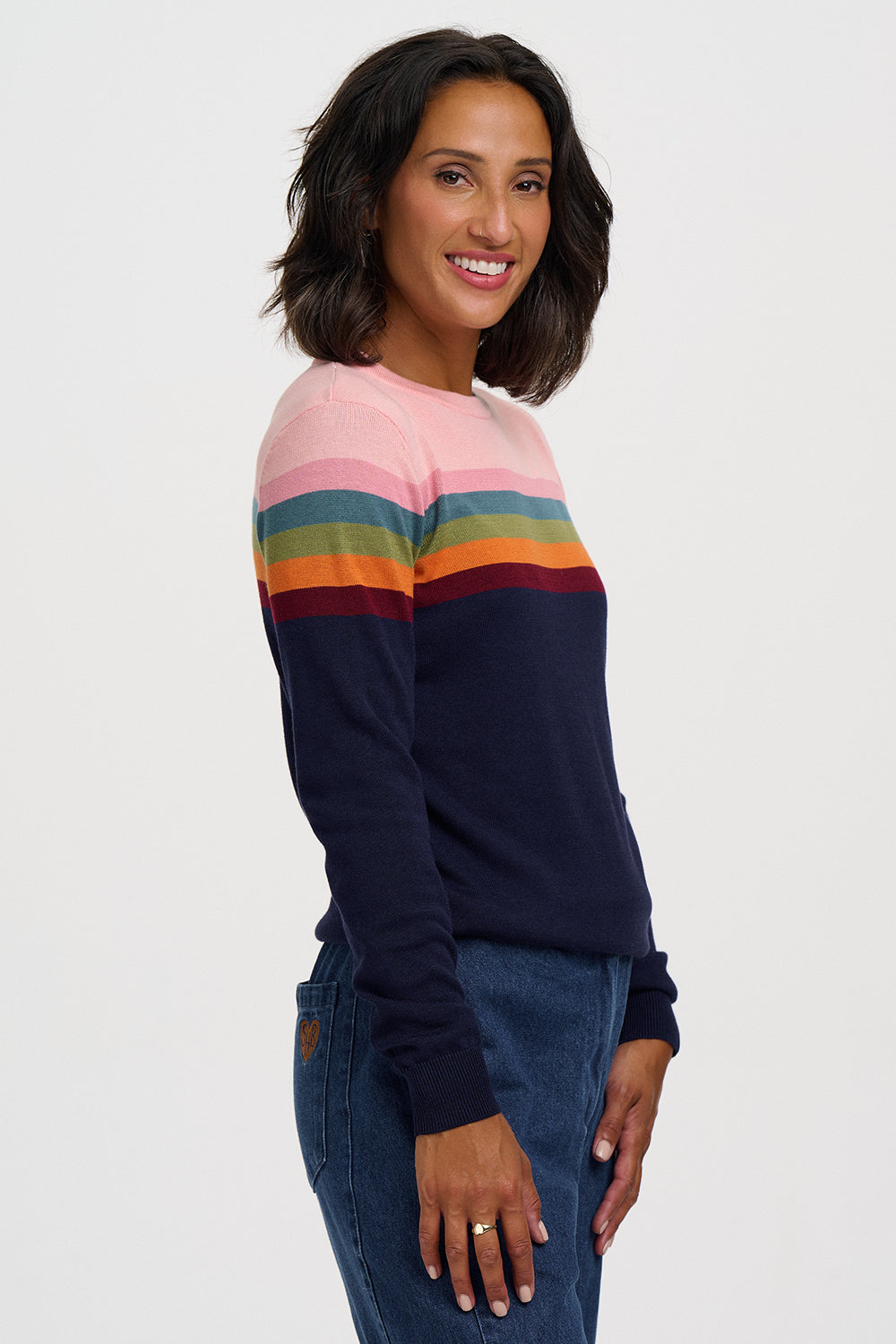 A woman wearing a long-sleeve crewneck sweater with horizontal rainbow stripes across the chest, paired with blue jeans, photographed against a light gray studio background.