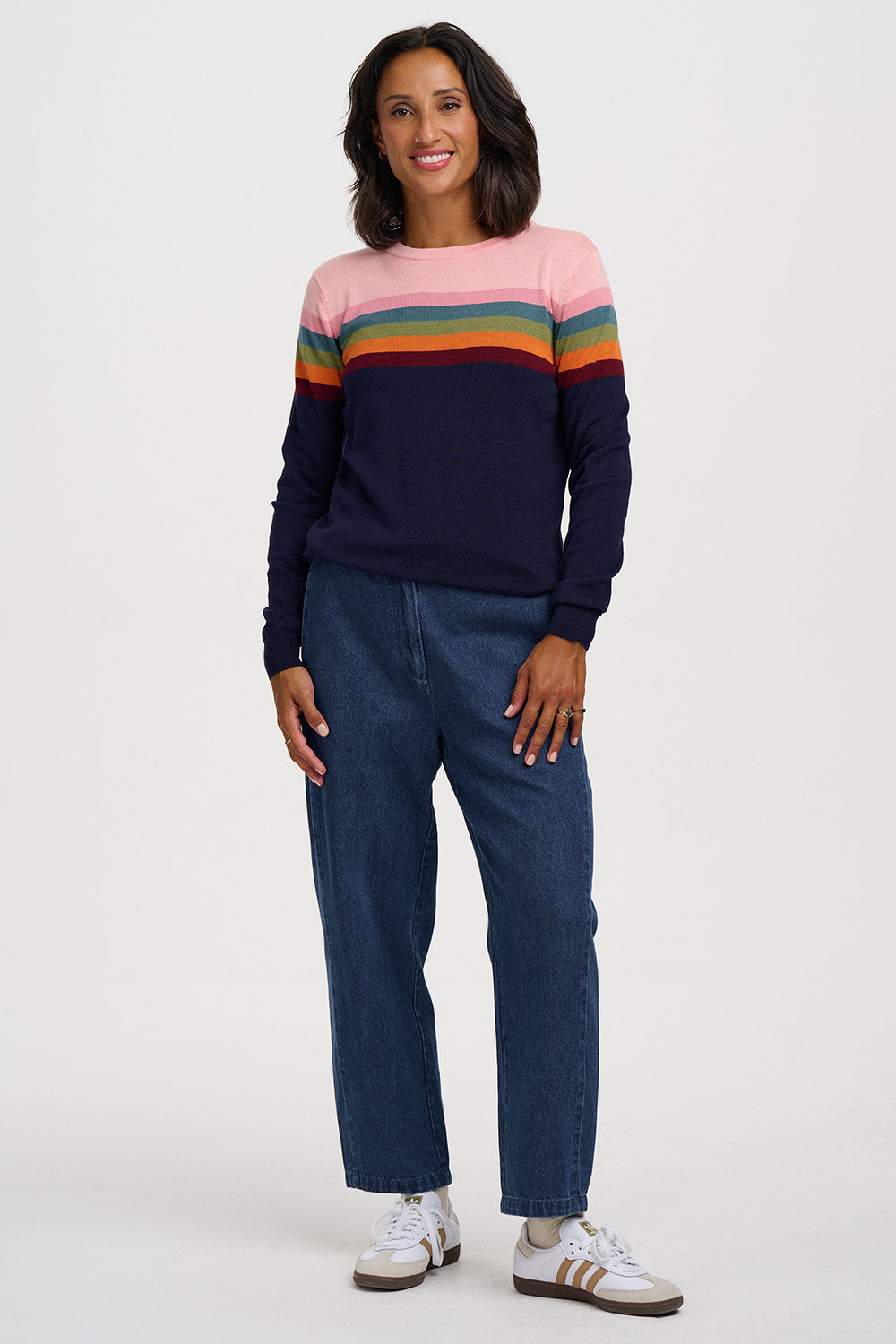 A woman modeling a long-sleeve crewneck sweater with horizontal stripes in pink, orange, yellow, green, and navy blue, paired with dark blue jeans and white sneakers, against a light gray studio background.
