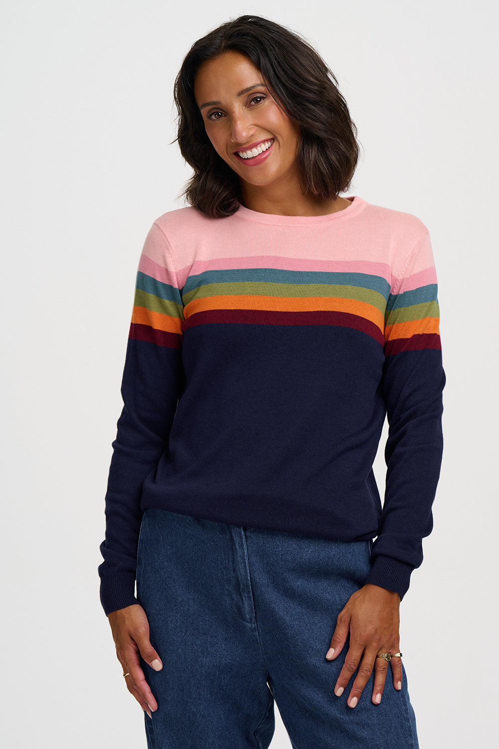 A woman wearing a long-sleeve crewneck sweater with horizontal rainbow stripes across the chest and upper arms, paired with dark blue jeans, against a light gray studio background.