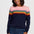 A woman wearing a long-sleeve crewneck sweater with horizontal rainbow stripes across the chest and upper arms, paired with dark blue jeans, against a light gray studio background.