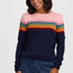 A woman wearing a long-sleeve crewneck sweater with horizontal rainbow stripes across the chest and upper arms, paired with dark blue jeans, against a light gray studio background.