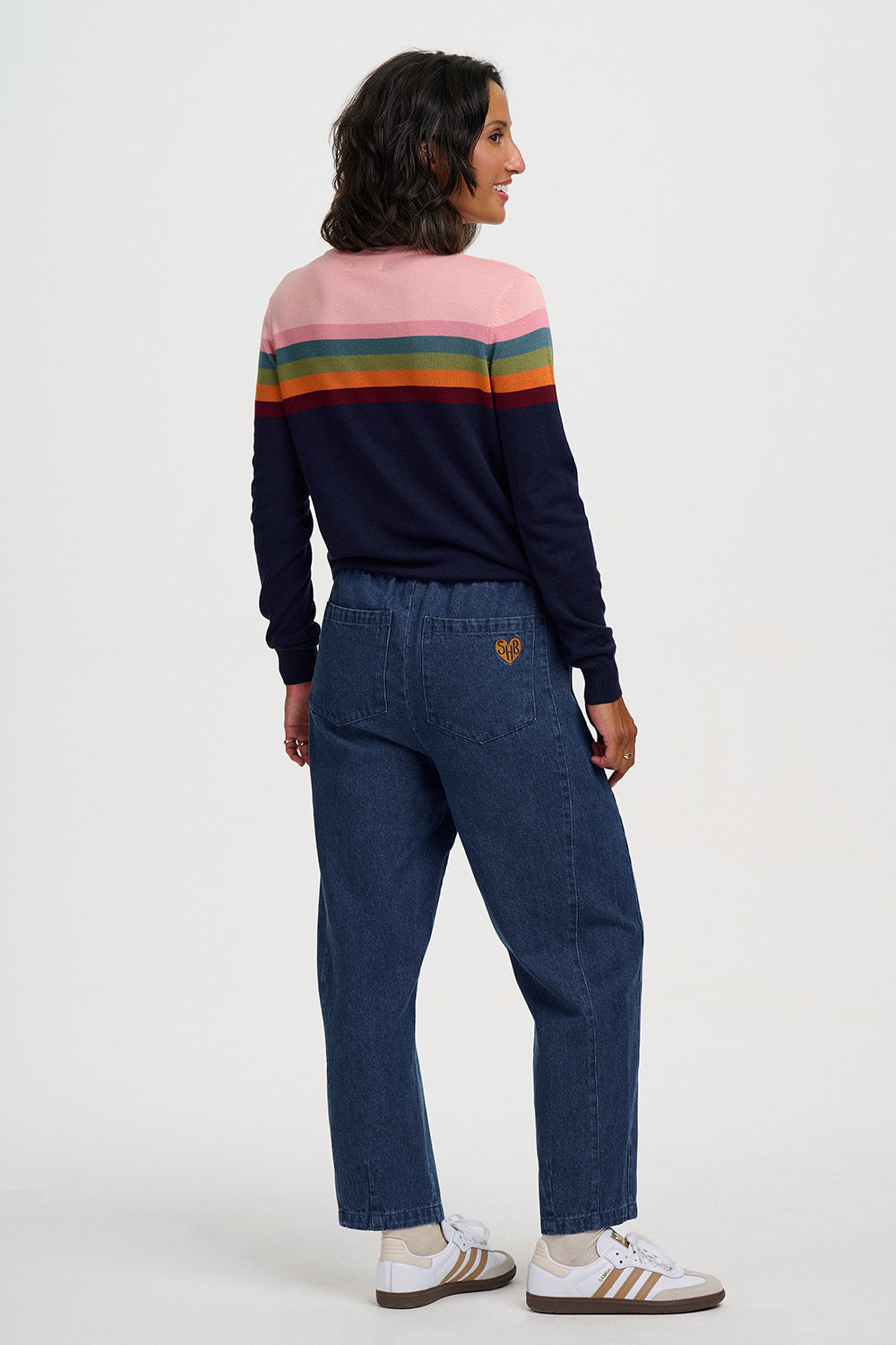 A woman is shown from the back wearing a navy long-sleeve sweater with a pink, green, orange, and yellow striped accent across the upper back, paired with dark blue jeans and white sneakers with gold stripes, against a plain white studio background.
