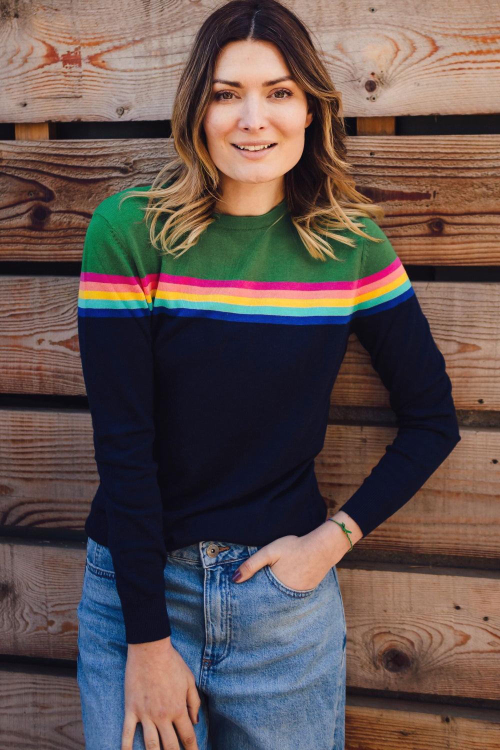 A woman wearing a navy and green color-blocked jumper with a rainbow split design across the chest, paired with blue jeans.
