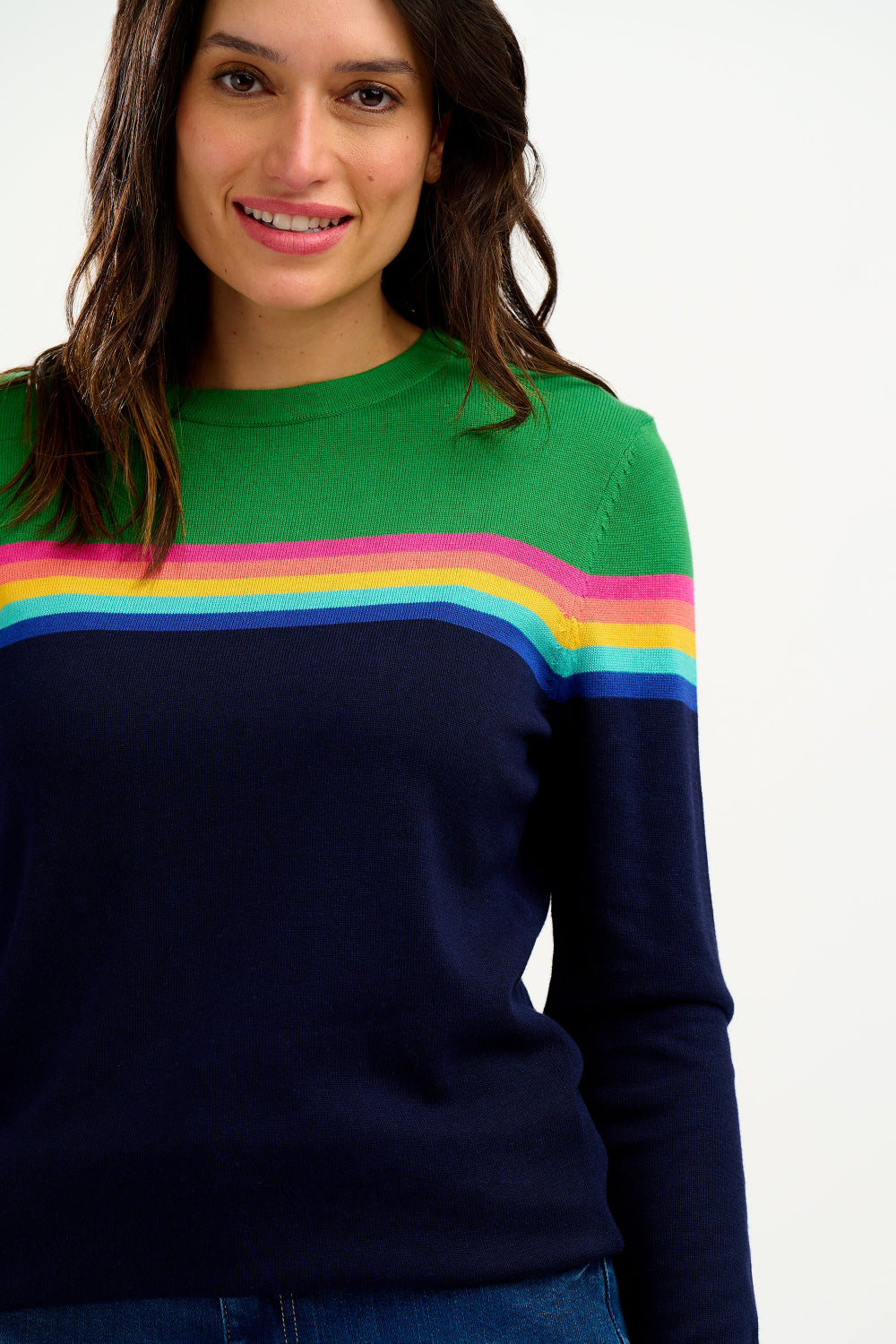 A woman wearing a navy and green color-blocked jumper with a rainbow split design across the chest, paired with blue jeans.