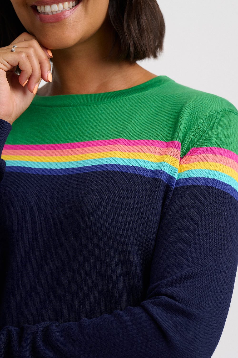 A close-up of a woman wearing a long-sleeve knit sweater with a green yoke, navy body, and horizontal rainbow stripes across the chest and shoulders.