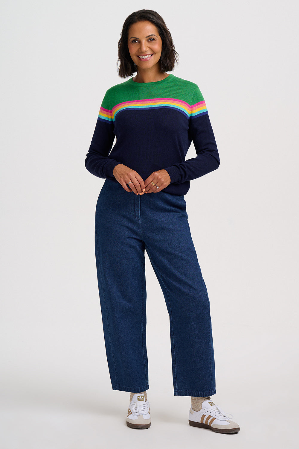 A woman modeling a navy blue long-sleeve knit sweater with a colorful striped yoke in green, pink, and orange, paired with wide-leg blue jeans and white sneakers, against a light gray studio background.