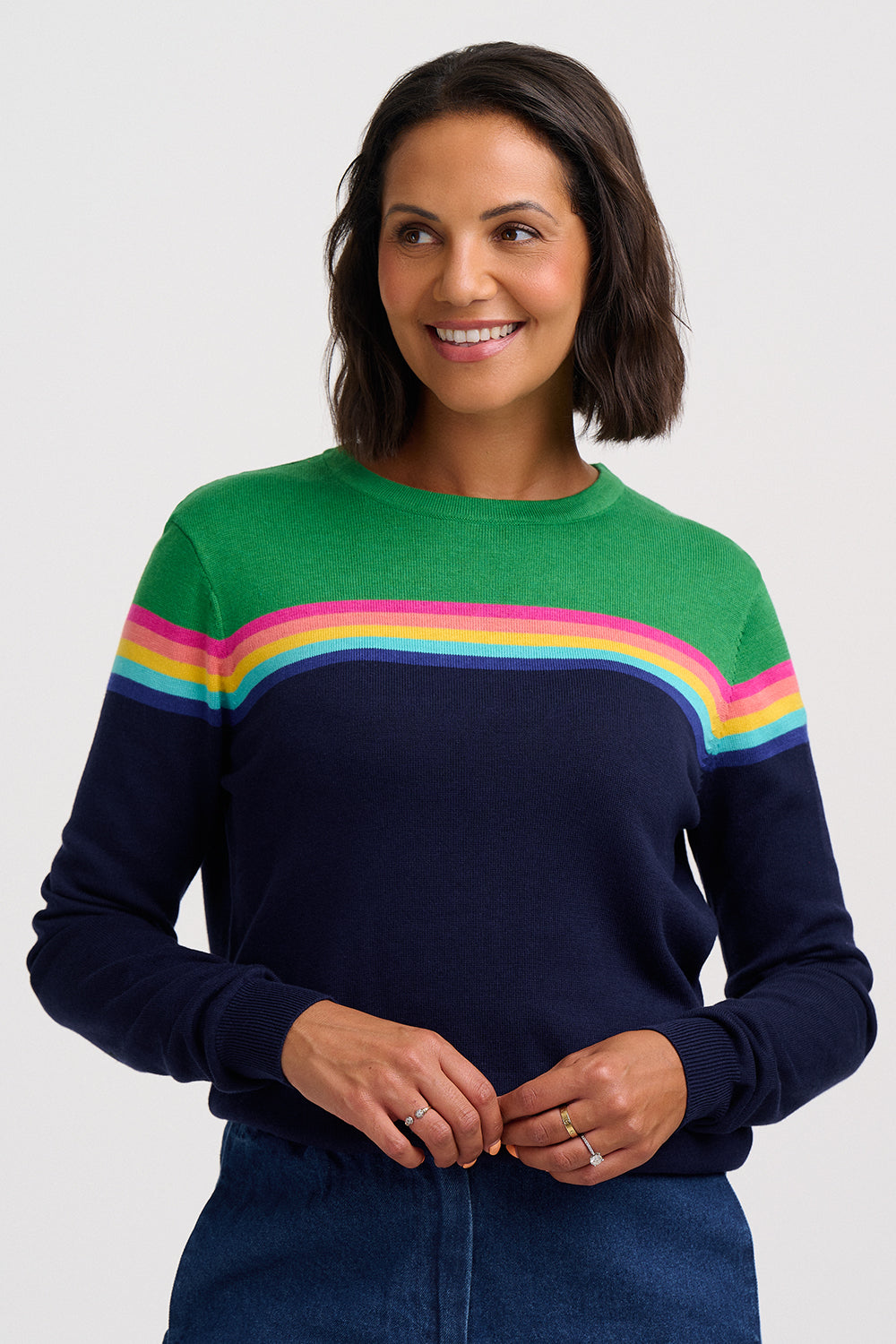 A woman wearing a crewneck sweater with a color-blocked design featuring green on the upper chest, a horizontal stripe of pink, yellow, and light blue across the middle, and navy blue on the lower body and sleeves, paired with dark blue jeans against a light gray background.