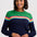 A woman wearing a crewneck sweater with a color-blocked design featuring green on the upper chest, a horizontal stripe of pink, yellow, and light blue across the middle, and navy blue on the lower body and sleeves, paired with dark blue jeans against a light gray background.