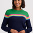 A woman wearing a crewneck sweater with a color-blocked design featuring green on the upper chest, a horizontal stripe of pink, yellow, and light blue across the middle, and navy blue on the lower body and sleeves, paired with dark blue jeans against a light gray background.
