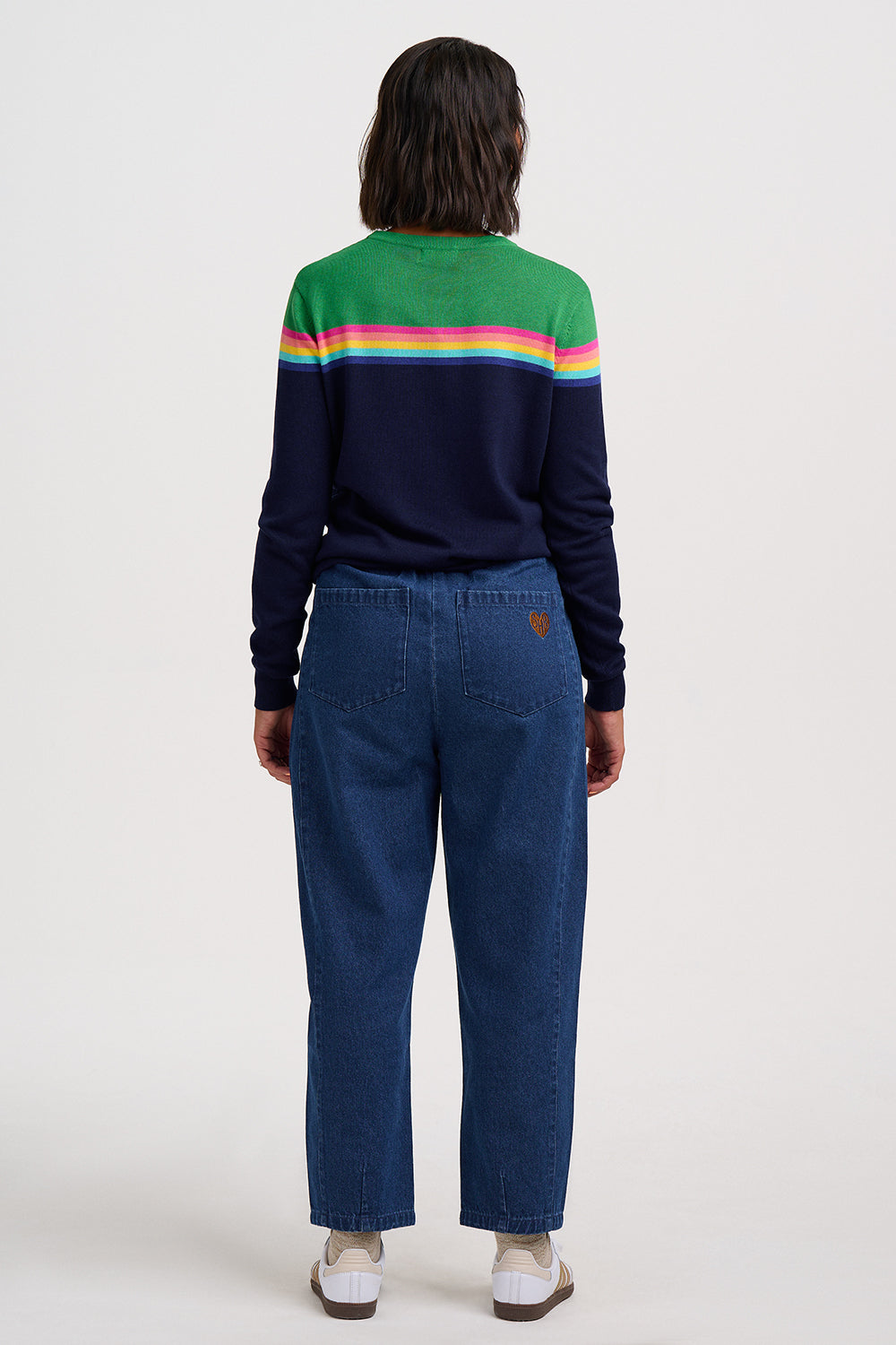 A model viewed from the back wearing a navy blue sweater with a green yoke and rainbow stripes across the shoulders, paired with blue jeans and white sneakers, against a plain light gray background.