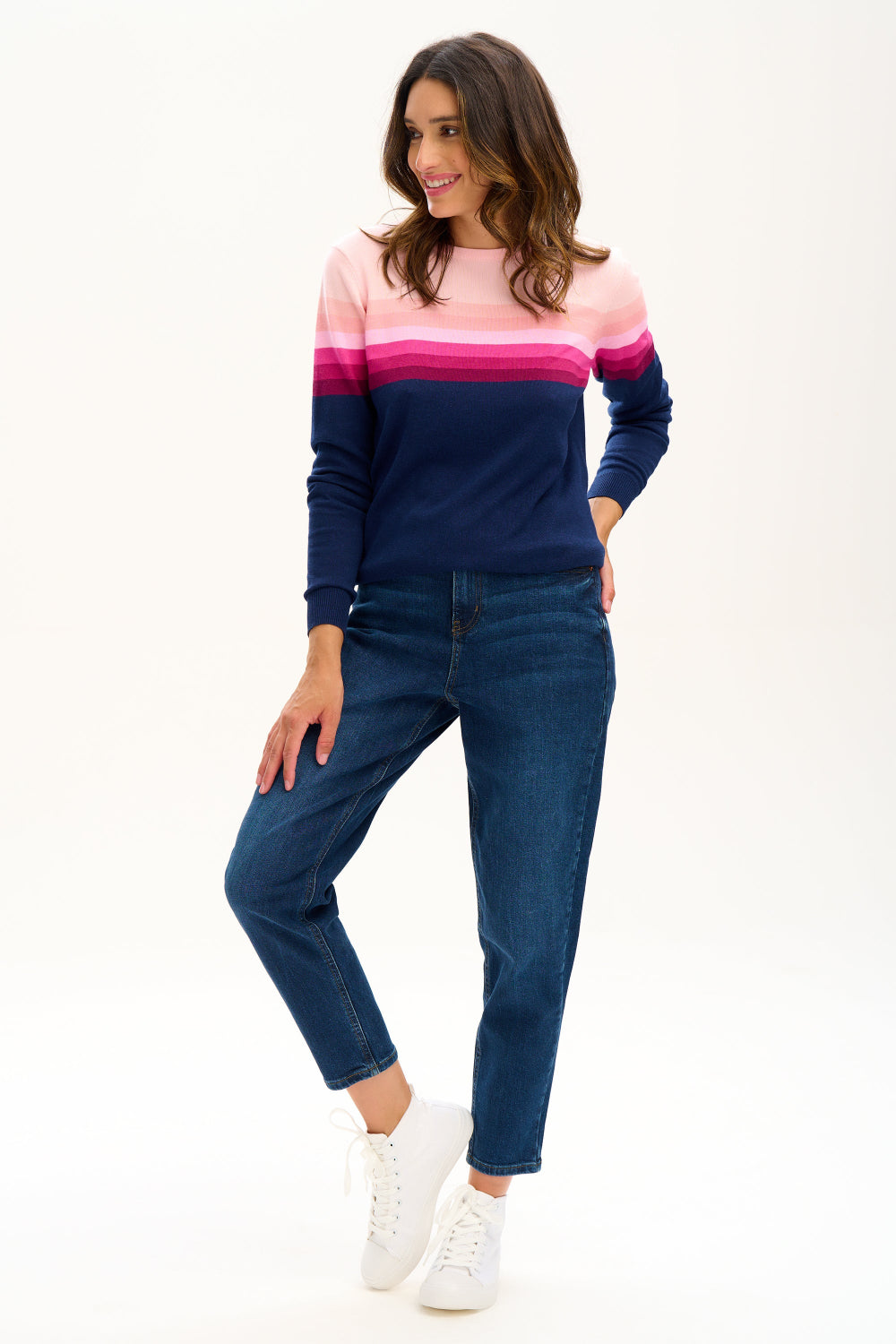 Sugarhill Brighton model wearing a navy jumper with pink gradient stripes, paired with jeans, standing against a white background.