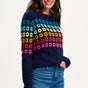 Sugarhill Brighton model wearing a navy blue jumper with a colourful geometric flower pattern, paired with jeans.