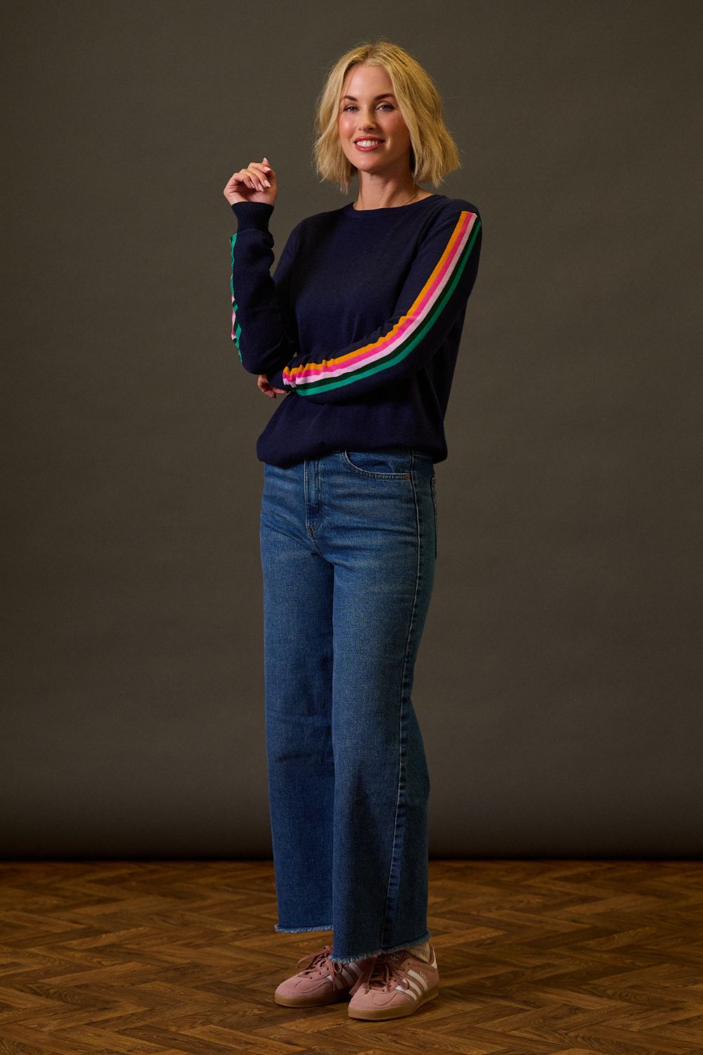 Woman wearing a navy sweater with colorful stripes and blue jeans against a dark background