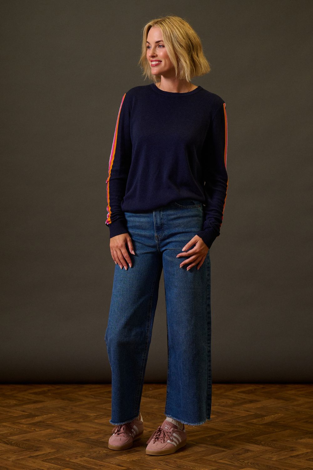 Woman wearing a navy sweater with orange accents and blue jeans against a dark background