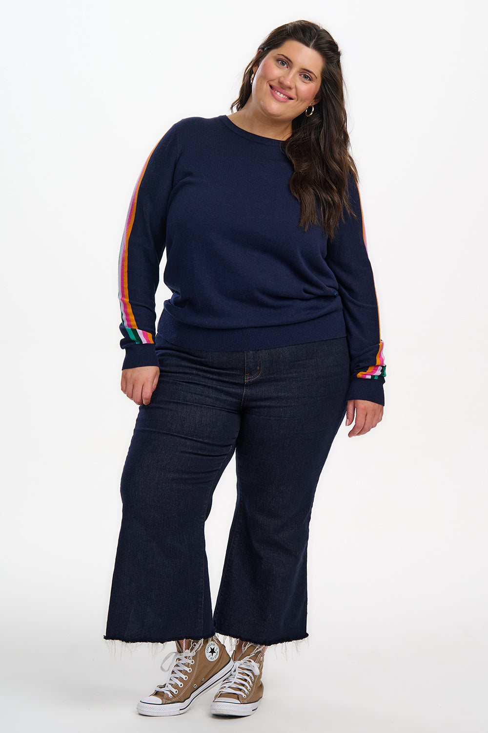 Woman wearing a navy sweatshirt with colorful sleeves and dark jeans on a white background