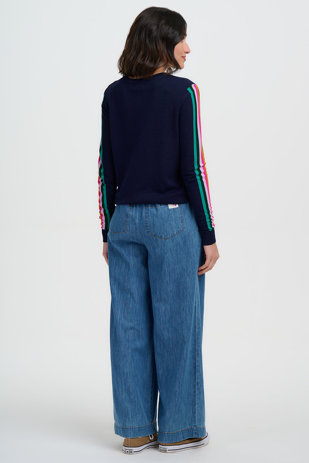 Person wearing a navy sweater with colorful stripes and blue jeans on a white background