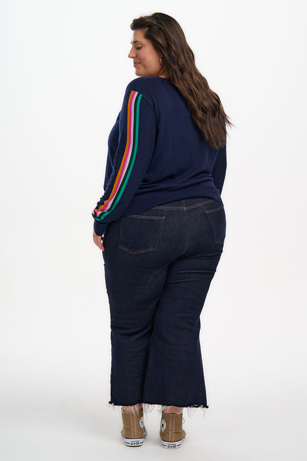 Person wearing a navy blue sweater with colorful stripes on the sleeves and dark jeans against a white background