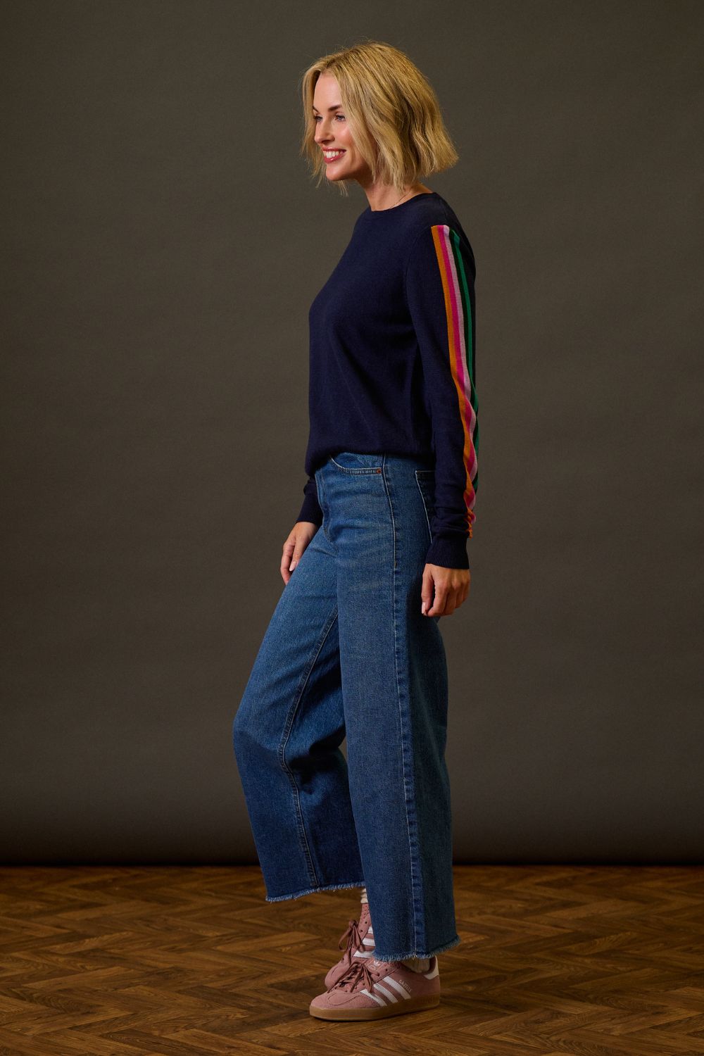 Woman wearing a navy sweater with colorful sleeves and blue jeans against a brown background