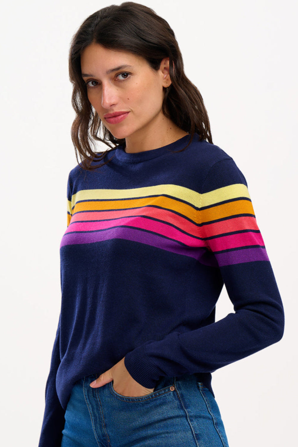 Sugarhill Brighton model wearing a navy blue jumper with horizontal retro stripe bands in pink, yellow, and other colours across the chest and shoulders.