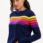 Sugarhill Brighton model wearing a navy blue jumper with horizontal retro stripe bands in pink, yellow, and other colours across the chest and shoulders.