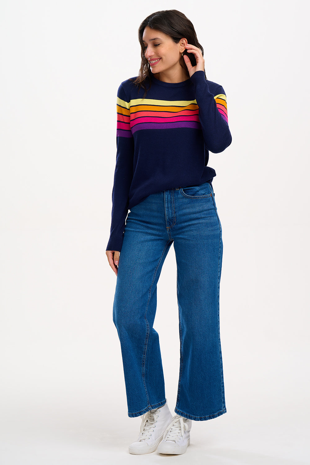 Sugarhill Brighton model wearing a navy blue jumper with horizontal retro stripe bands in pink, yellow, and other colours across the chest and shoulders.
