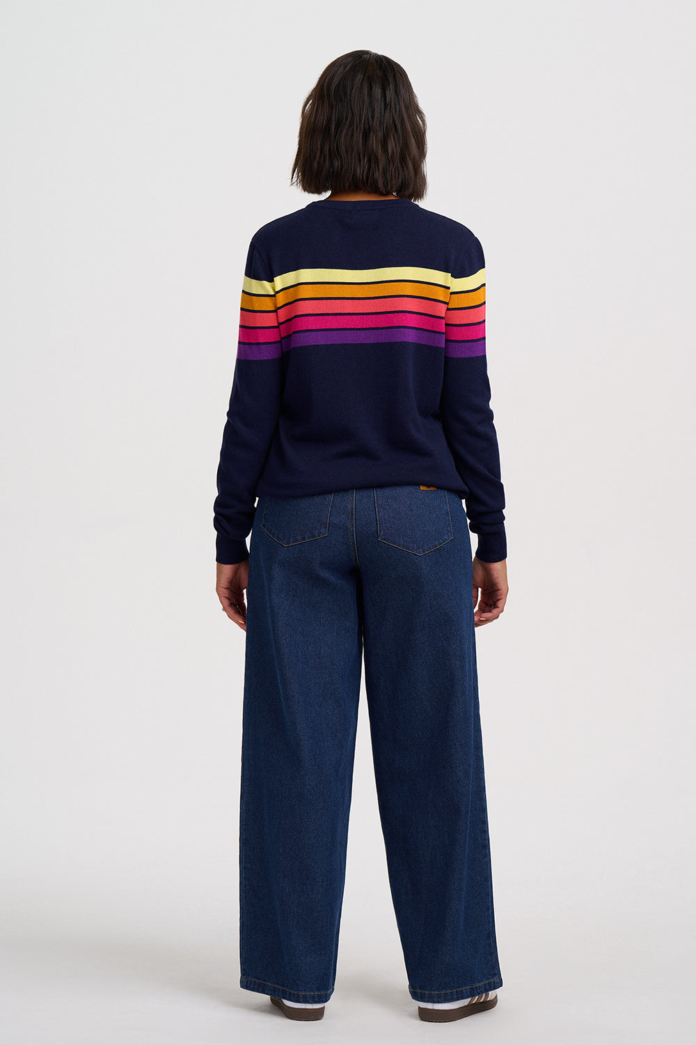 A person shown from the back wearing a navy sweater with horizontal yellow, orange, pink, and purple stripes across the upper back and wide-leg dark blue jeans, photographed against a plain light gray studio background.