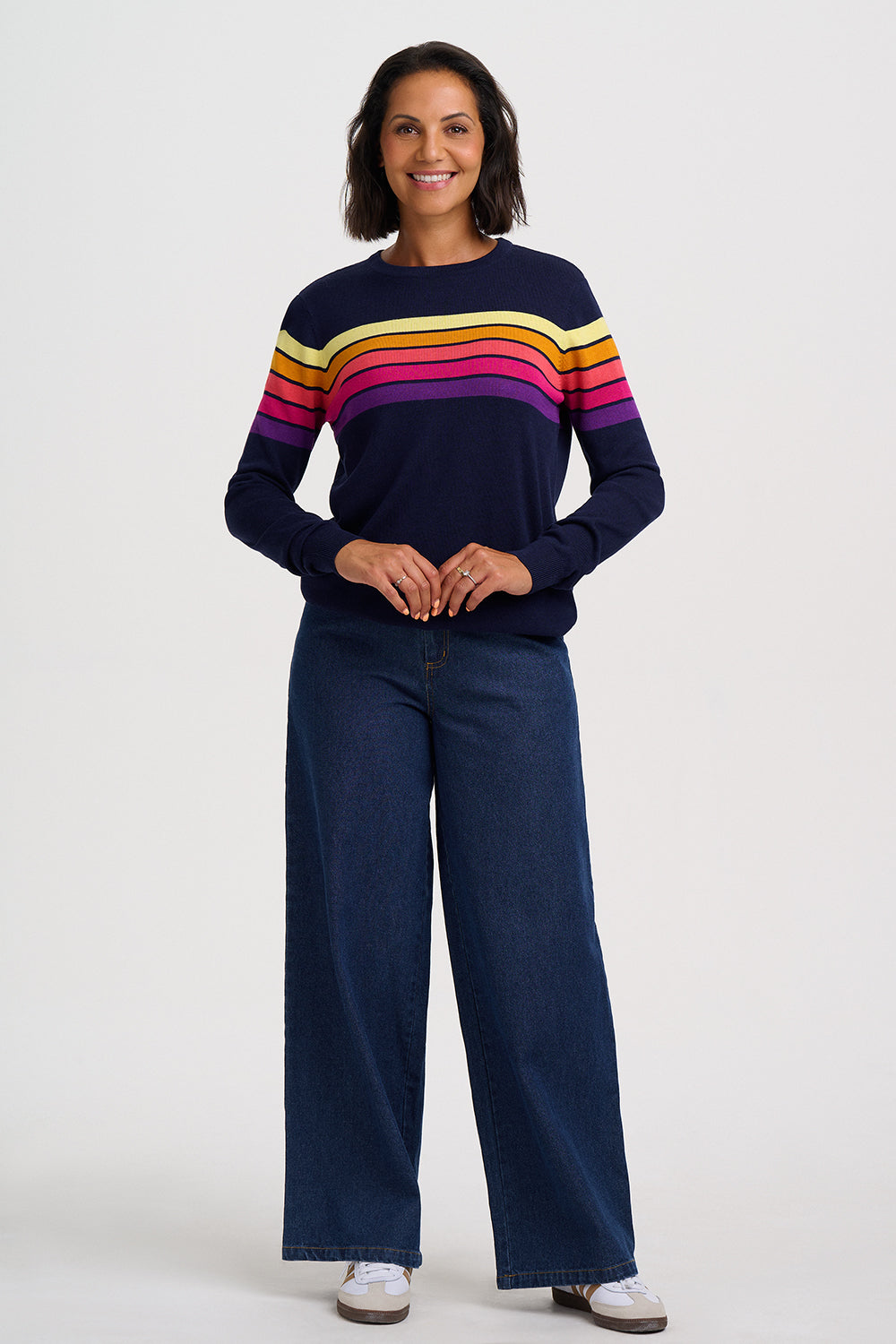 A woman modeling a navy blue long-sleeve knit sweater with horizontal rainbow stripes across the chest and shoulders, paired with dark wide-leg jeans and white sneakers, against a light gray studio background.