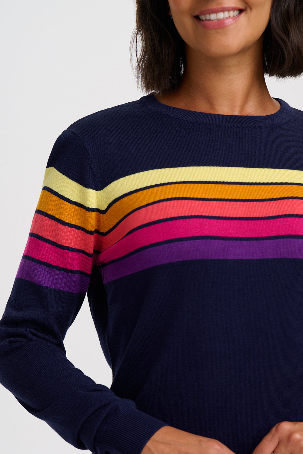 A close-up of a woman wearing a navy blue crewneck sweater with horizontal rainbow stripes across the chest and shoulders in yellow, orange, pink, and purple, against a light gray background.