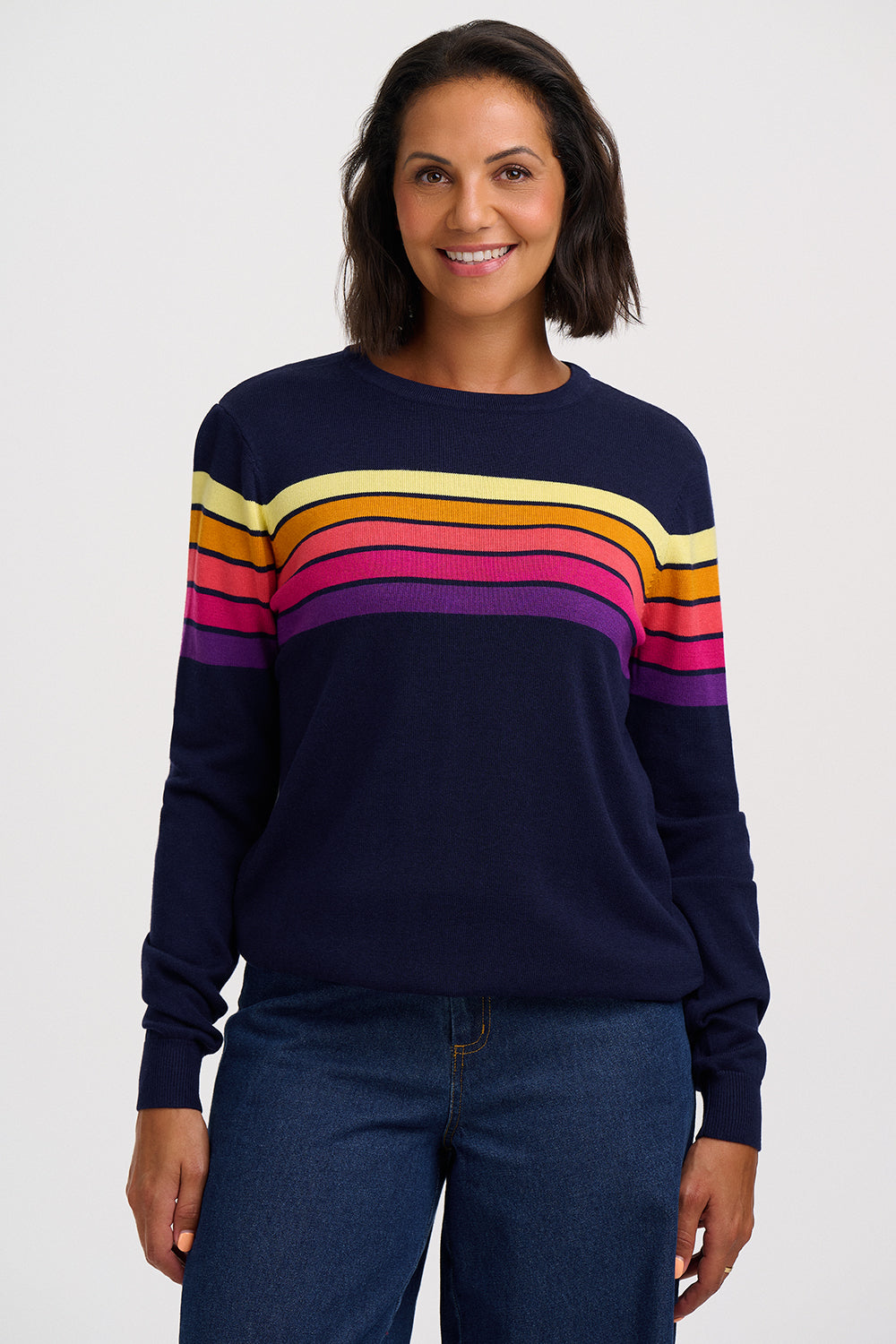 A woman wearing a navy blue crewneck sweater with horizontal rainbow stripes across the chest and upper arms, paired with dark blue jeans, against a light gray studio background.