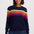 A woman wearing a navy blue crewneck sweater with horizontal rainbow stripes across the chest and upper arms, paired with dark blue jeans, against a light gray studio background.