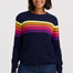 A woman wearing a navy blue crewneck sweater with horizontal rainbow stripes across the chest and upper arms, paired with dark blue jeans, against a light gray studio background.