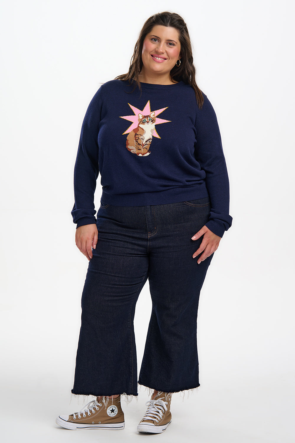 Woman wearing a navy blue sweatshirt with a graphic design, dark jeans, and brown sneakers on a white background