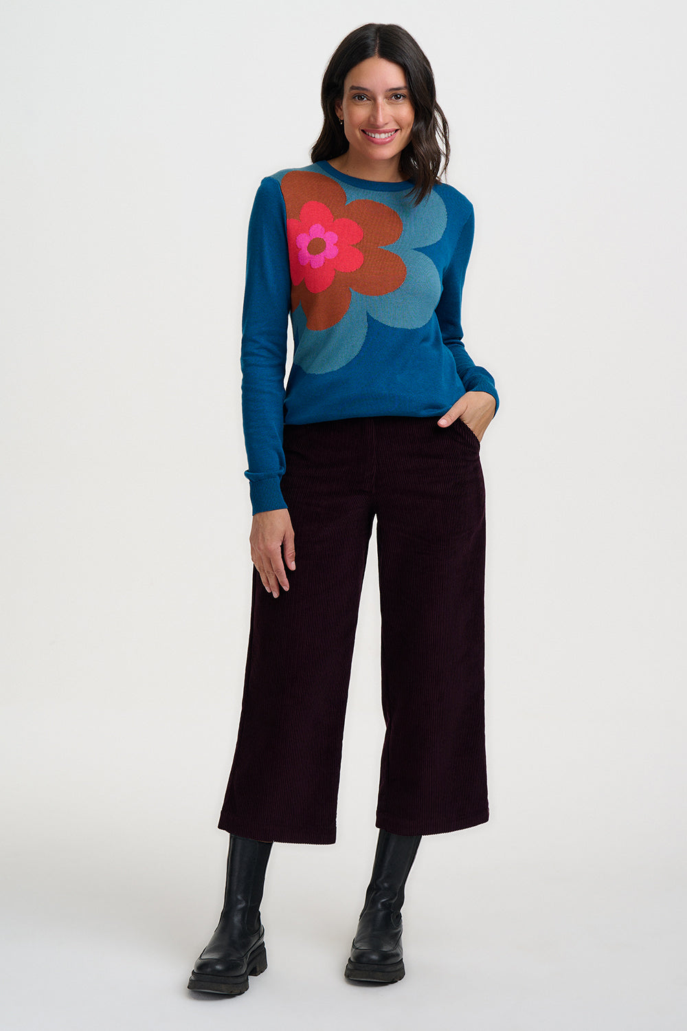 Woman wearing a blue sweater with a colorful floral design, black pants, and black boots on a white background