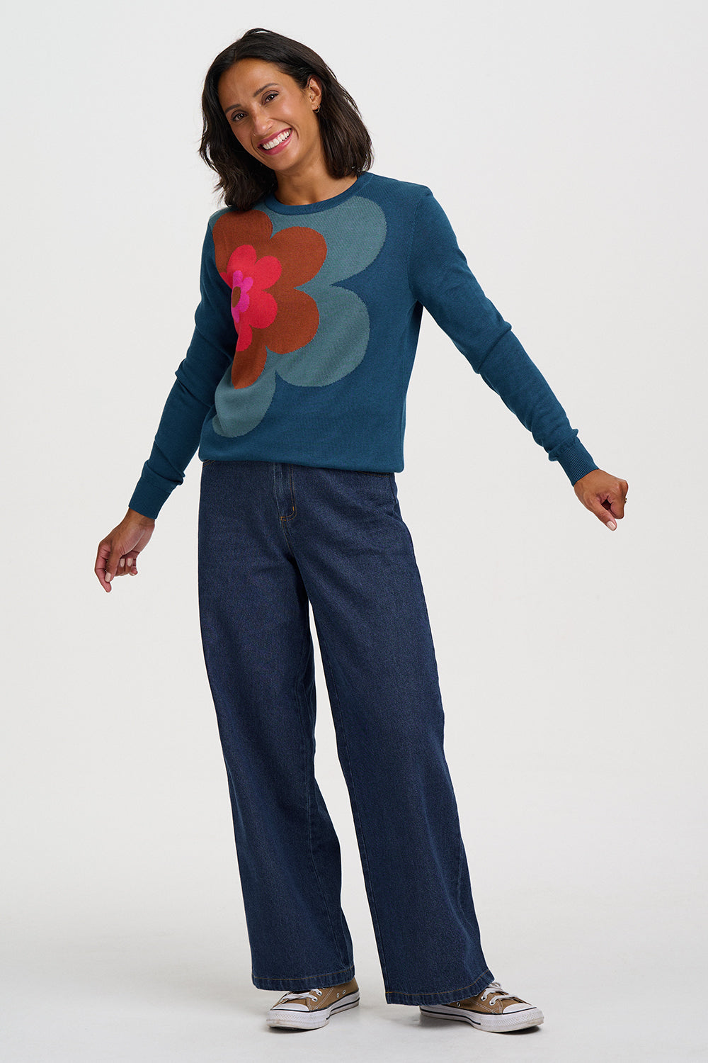 A woman modeling a teal blue long-sleeve knit sweater with a large abstract floral pattern in pink, red, and blue-gray on the front, paired with dark wide-leg jeans and canvas sneakers against a light gray studio background.